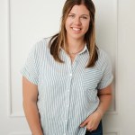 A smiling woman with long brown hair wearing a light blue and white striped button-up shirt, posing against a neutral background.