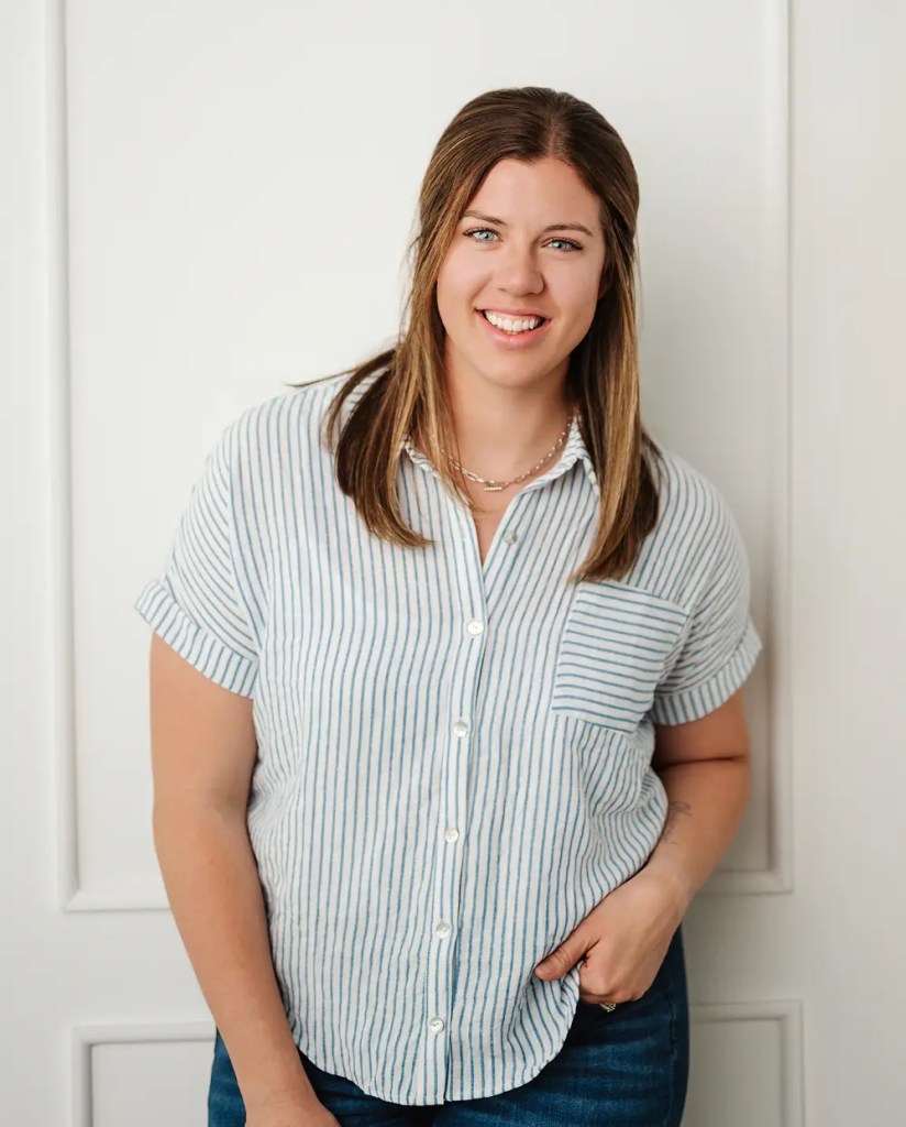 A smiling woman with long, straight hair is wearing a striped button-up shirt and standing against a plain white wall.